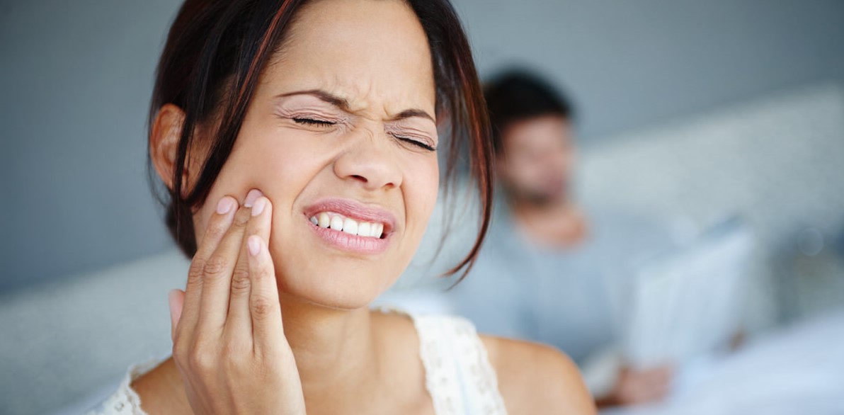 Shot of a woman sitting on the side of her bed with bad toothache with her boyfriend in the background